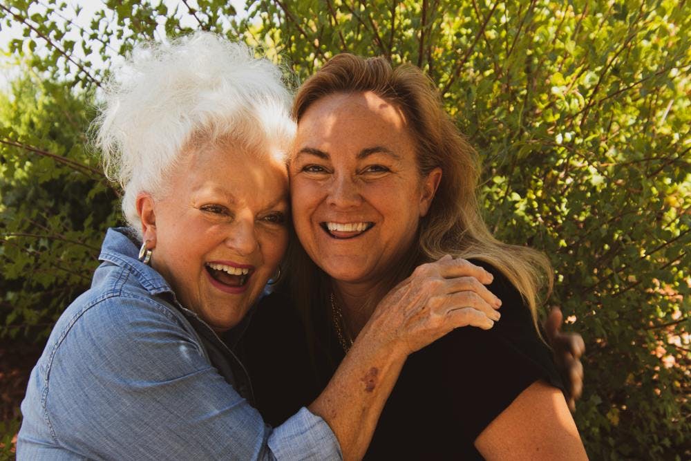 An older, white-haired woman wearing a blue button-up smiling and hugging an older, smiling woman wearing a black t-shirt. 